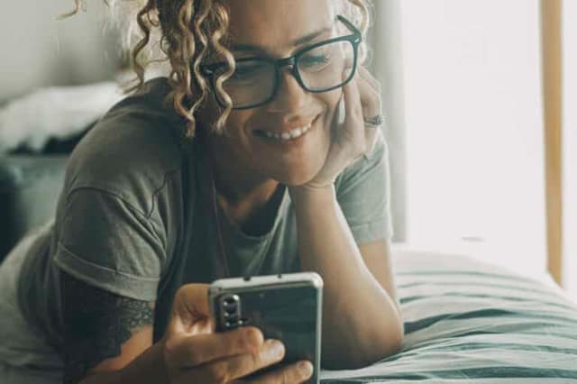 Portrait d'une femme heureuse et joyeuse, allongée sur le lit et utilisant un téléphone portable pour discuter et regarder des contenus en ligne. Des femmes modernes avec un smartphone. Bavarder avec des amis.